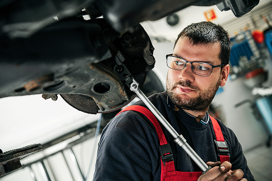 Ein erfahrener Mechaniker mit Brille und rotem Overall führt in der hochmodernen Werkstatt der Frey Auto AG eine MFK-Inspektion und einen Reifenservice an der Unterseite eines Toyota-Fahrzeugs durch. Die Werkstatt ist auf umfassende Autoservices spezialisiert, darunter Karosseriearbeiten und Wartung für Topmarken wie Lexus und Suzuki, um Spitzenleistung zu gewährleisten.
