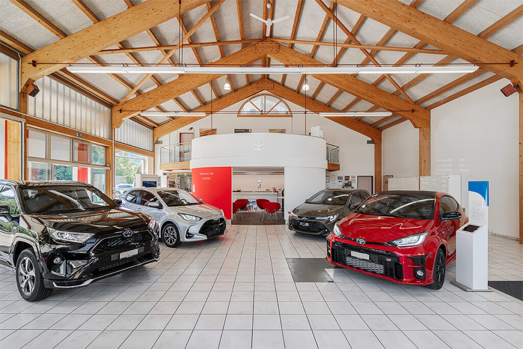 Der Ausstellungsraum der Frey Auto AG bietet eine Auswahl an Fahrzeugen der Marken Toyota, Lexus und Suzuki in Schwarz, Silber und Rot, die auf einem polierten Fliesenboden ausgestellt sind. Der Raum wird durch Holzbalken und große Fenster akzentuiert, die für eine bessere natürliche Beleuchtung sorgen und den makellosen Zustand jedes Autos zur Schau stellen. Frey Auto AG ist auf umfassende Autodienstleistungen spezialisiert, darunter MFK-Inspektionen, detaillierter Reifenservice, professionelle Karosserielösungen, sorgfältige Autowäschen und zuverlässiger Autoverkauf, der auf die Kundenbedürfnisse zugeschnitten ist.
