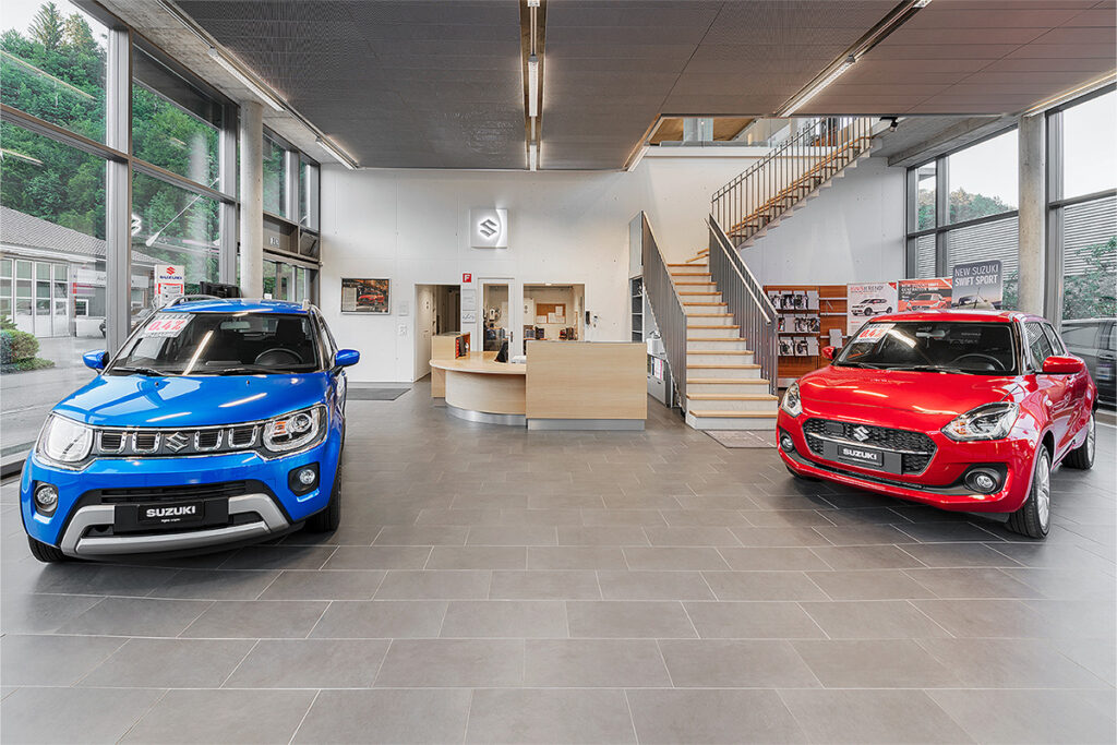 Entdecken Sie den Showroom der Frey Auto AG, in dem unsere außergewöhnlichen Suzuki-Modelle im Mittelpunkt stehen: ein auffälliges blaues SUV und ein leuchtend rotes Fließheck. Erleben Sie unsere umfassenden Kfz-Dienstleistungen, darunter fachmännische MFK-Inspektionen, zuverlässiger Reifenservice, präzise Karosserielösungen, professioneller Autoverkauf für Toyota-, Lexus- und Suzuki-Modelle sowie sorgfältige Autowaschoptionen. Erfahren Sie mehr an unserem einladenden Empfangstresen oder wagen Sie sich weiter durch die elegante Treppe, die Ihnen weitere Einblicke in die Angebote von Toyota und Lexus bietet.