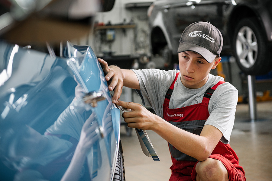 Ein junger Mechaniker in rotem Overall und grauem Hemd führt in der Werkstatt der Frey Auto AG, die auf MFK-Inspektionen spezialisiert ist, sorgfältig Wartungsarbeiten an einem blauen Toyota durch. Die gut ausgestattete Werkstatt bietet umfassende Kfz-Dienstleistungen an, darunter Reifenwechsel und Achsvermessung, präzise Karosseriereparaturen, gründliche Autowäschen und spezielle Suzuki-Wartung. Auch für die Detaillierung von Lexus-Fahrzeugen ist fachmännische Aufmerksamkeit verfügbar.
