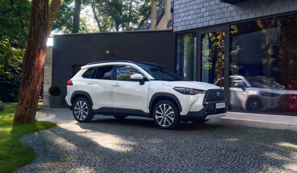Ein weißer Toyota-SUV parkt auf der Kopfsteinpflasterauffahrt neben dem modernen Gebäude der Frey Auto AG mit seinen großen Glasfenstern. Das Autohaus bietet umfassende Kfz-Dienstleistungen an, darunter MFK-Inspektion, Reifenservice, Karosseriearbeiten, Autowäsche und autorisierten Autoverkauf von Topmarken wie Toyota, Lexus und Suzuki. Üppige Bäume umgeben den Bereich und unterstreichen die elegante Ästhetik.