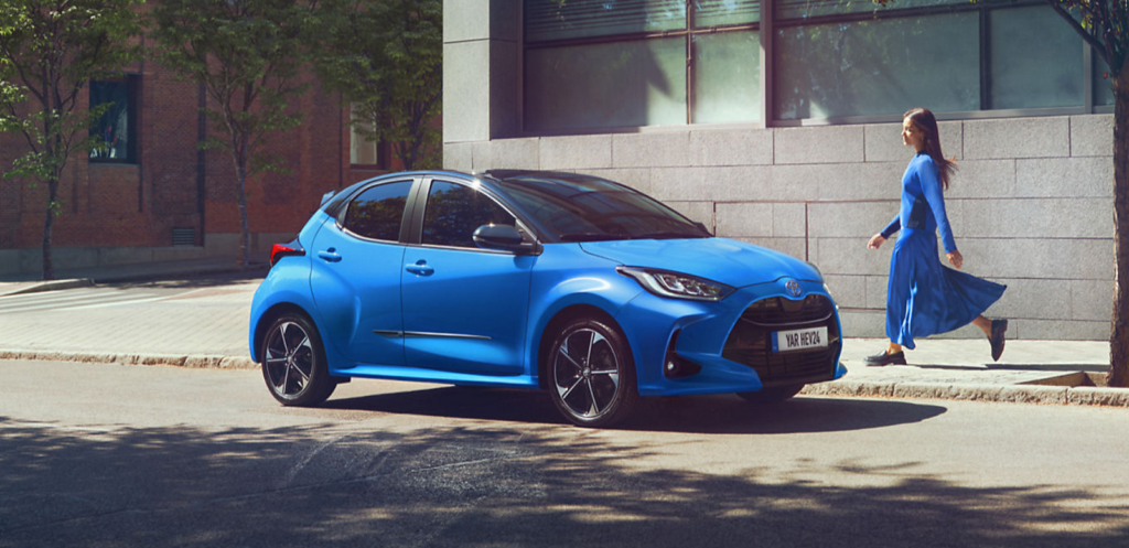 Eine blaue Toyota-Limousine steht auf einer Stadtstraße in der Nähe eines eleganten, modernen Gebäudes. Eine Person in einem blauen Kleid läuft neben dem Auto auf dem Bürgersteig entlang. Frey Auto AG bietet umfassende Dienstleistungen für Toyota-, Lexus- und Suzuki-Fahrzeuge an, darunter MFK-Inspektion, Reifenservice, routinemäßige Wartungsdienste, fachmännische Karosseriereparaturen und professionellen Autoverkauf. Besuchen Sie uns für außergewöhnliche Pflege in unserer hochmodernen Anlage, die auch bequeme Autowaschmöglichkeiten bietet, um Ihr Fahrzeug in Topzustand zu halten.