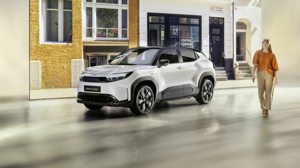 Ein weißer Toyota Urban Cruiser parkt vor dem Gebäude der Frey Auto AG, die auf MFK-Inspektionen, Reifenservice, allgemeine Fahrzeugwartung, Karosseriereparaturen und umfassenden Autoverkauf spezialisiert ist. Eine Frau in einem orangefarbenen Oberteil und beigen Hosen geht in der Nähe des Autohauses vorbei, das eine große Auswahl an Fahrzeugen von Topmarken wie Toyota, Lexus und Suzuki anbietet.