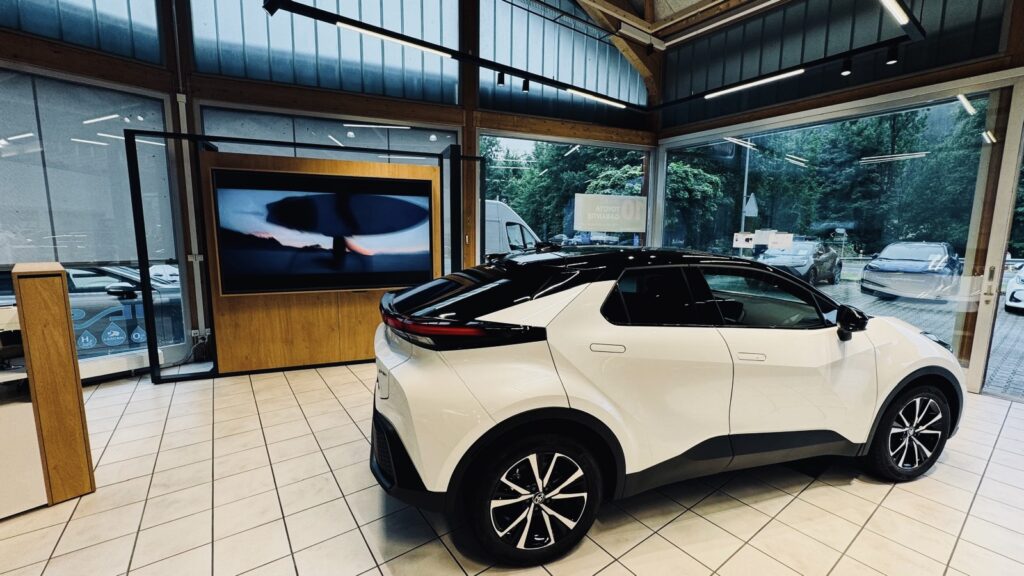 Ein weiss-schwarzer, moderner Toyota SUV im Showroom der Frey Auto AG, mit grossen Fenstern, gefliestem Boden und einem Bildschirm, auf dem ein Werbevideo läuft. Um das Fahrzeug herum sind Modelle von Lexus und Suzuki ausgestellt. Sichtbare Schilder weisen auf die Dienstleistungen der Frey Auto AG hin, darunter MFK-Inspektion, Reifenservice, Wartung, Karosseriereparaturen, zertifizierter Autoverkauf für Toyota-, Lexus- und Suzuki-Fahrzeuge sowie eine Autowaschanlage vor Ort.