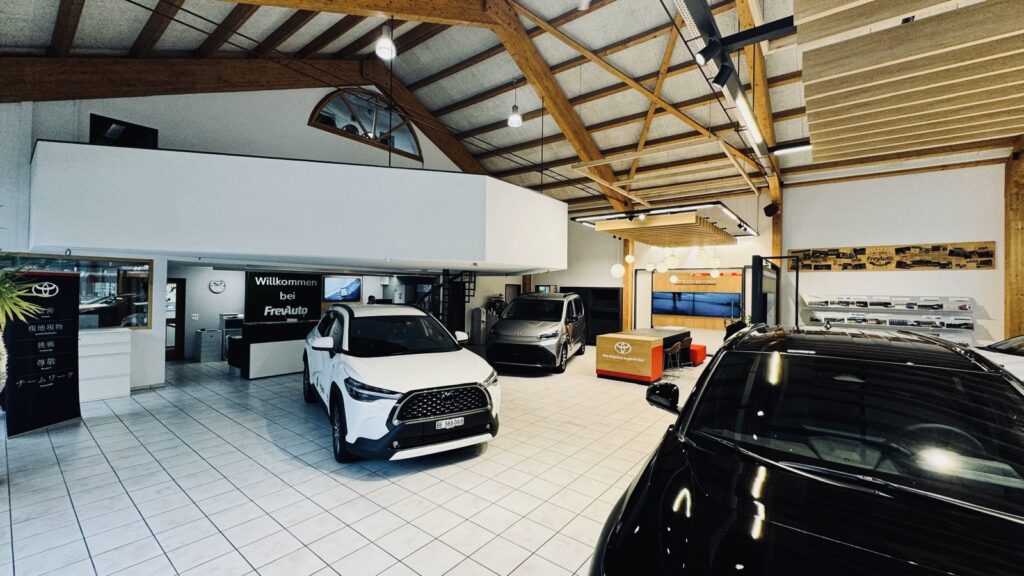 Modernes Autohaus der Frey Auto AG mit ausgestellten Fahrzeugen der Marken Toyota, Lexus und Suzuki; heller weisser Fliesenboden mit sichtbaren Holzbalken und einem deutschen "Willkommen"-Schild an der Rückwand. Sichtbare Bereiche sind die MFK-Inspektion, der professionelle Reifenservice, der Reparaturservice, die Karosserie-Spezialreparaturen, die zertifizierte Autoverkaufsberatung und die hauseigene Autowaschanlage, in der umfassende Automobillösungen präsentiert werden.