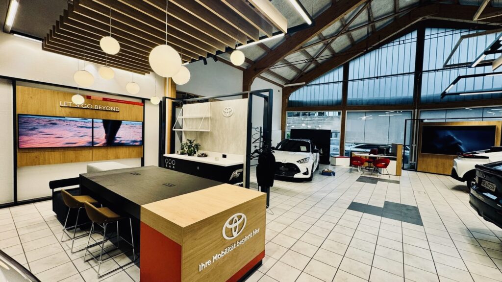 Moderner Toyota-Showroom bei Frey Auto AG mit ausgestellten Toyota-, Lexus- und Suzuki-Neufahrzeugen; großzügige Raumaufteilung mit großen Fenstern für natürliches Licht; eigener Empfangsschalter für Kundendienstanfragen wie MFK-Inspektionstermine, Reifenservicebuchungen und Karosserieberatungen; komfortabler Lounge-Bereich für wartende Kunden; digitale Bildschirme mit aktuellen Verkaufsangeboten, Wartungsdiensten, Autowaschaktionen und verfügbaren Originalteilen.