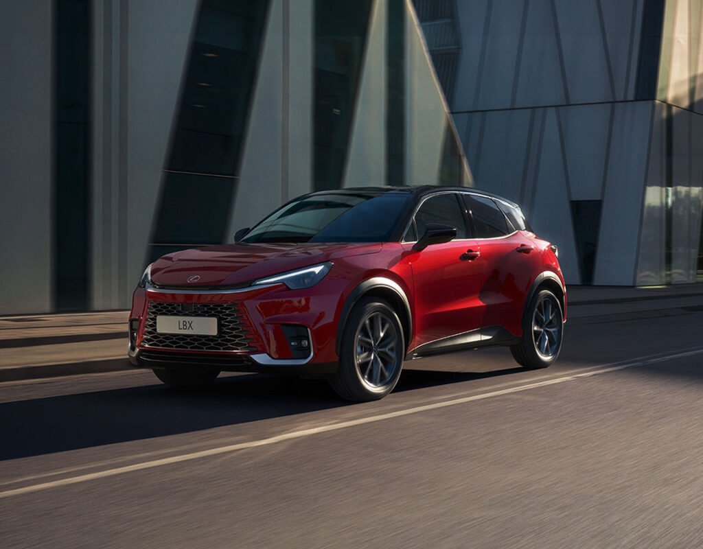 Ein roter Lexus LBX fährt auf einer Straße vor einem modernen Glasgebäude – ein Symbol für den Verkauf von Premium-Fahrzeugen und die Kompetenz im Kfz-Service der Frey Auto AG, die sich auf die Wartung von Toyota-, Lexus- und Suzuki-Fahrzeugen spezialisiert hat, einschließlich MFK-Prüfungen, Karosseriearbeiten und Reifenservice.