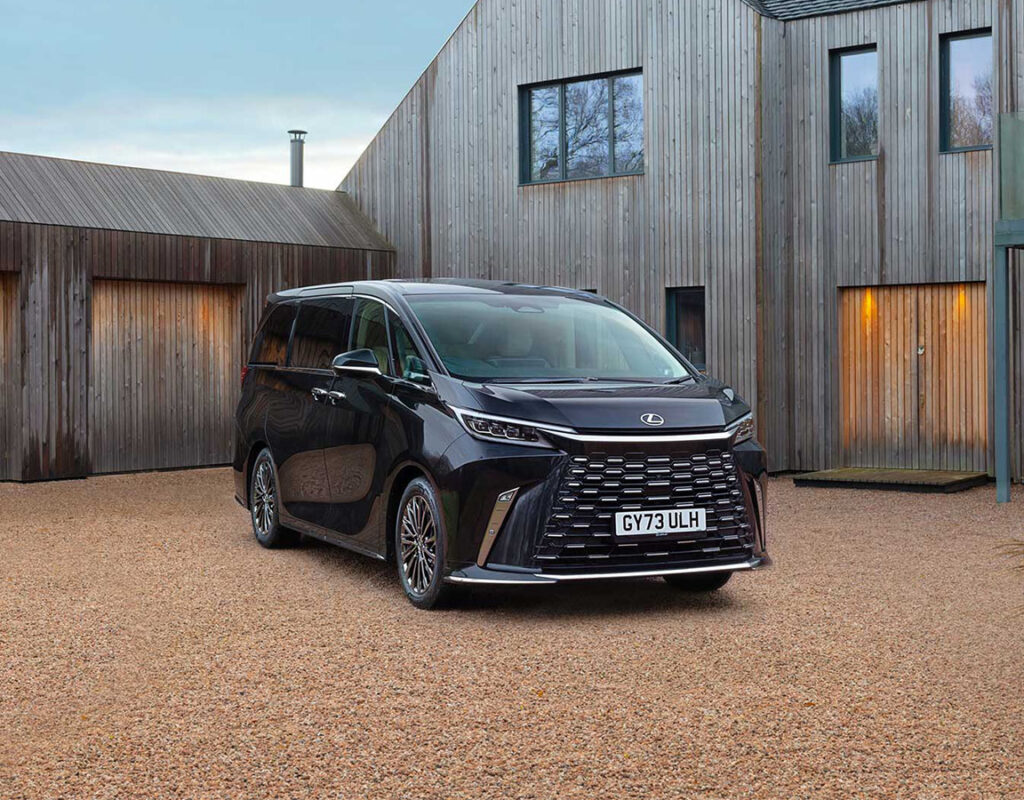 Ein schwarzer Lexus-Minivan, der auf einer Schotterauffahrt bei der Frey Auto AG geparkt ist, einem Fachbetrieb für Autoverkauf, Service und Karosseriearbeiten vor einem modernen Holzgebäude.