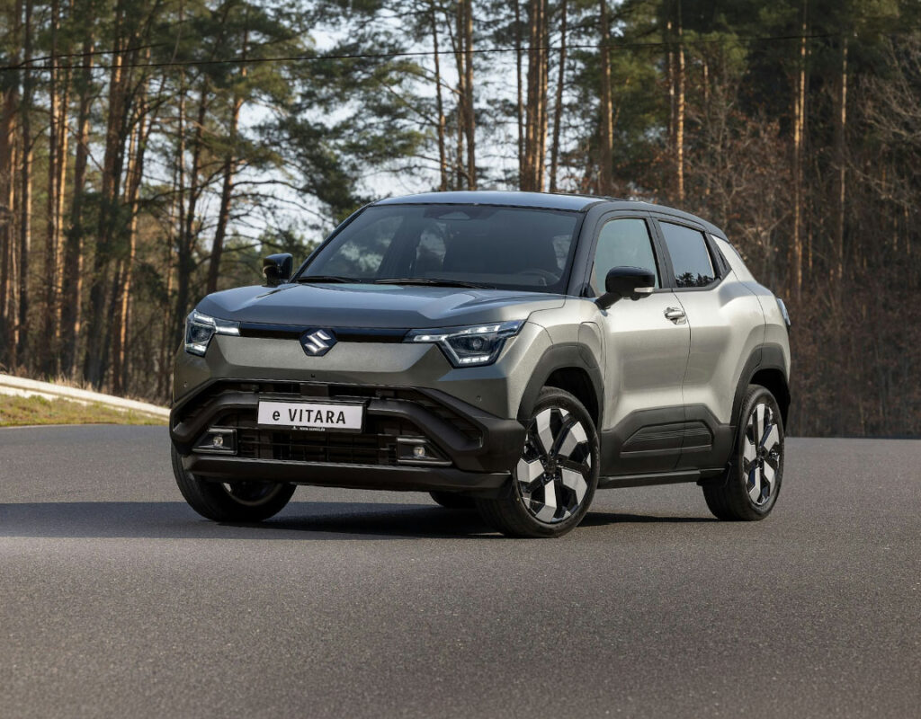Grauer Suzuki e-Vitara SUV auf einer Asphaltstraße vor einem Wald, erhältlich bei Frey Auto AG für den professionellen Verkauf von Suzuki-Fahrzeugen, Service, MFK-Prüfung, Reifenservice und Karosseriearbeiten.
