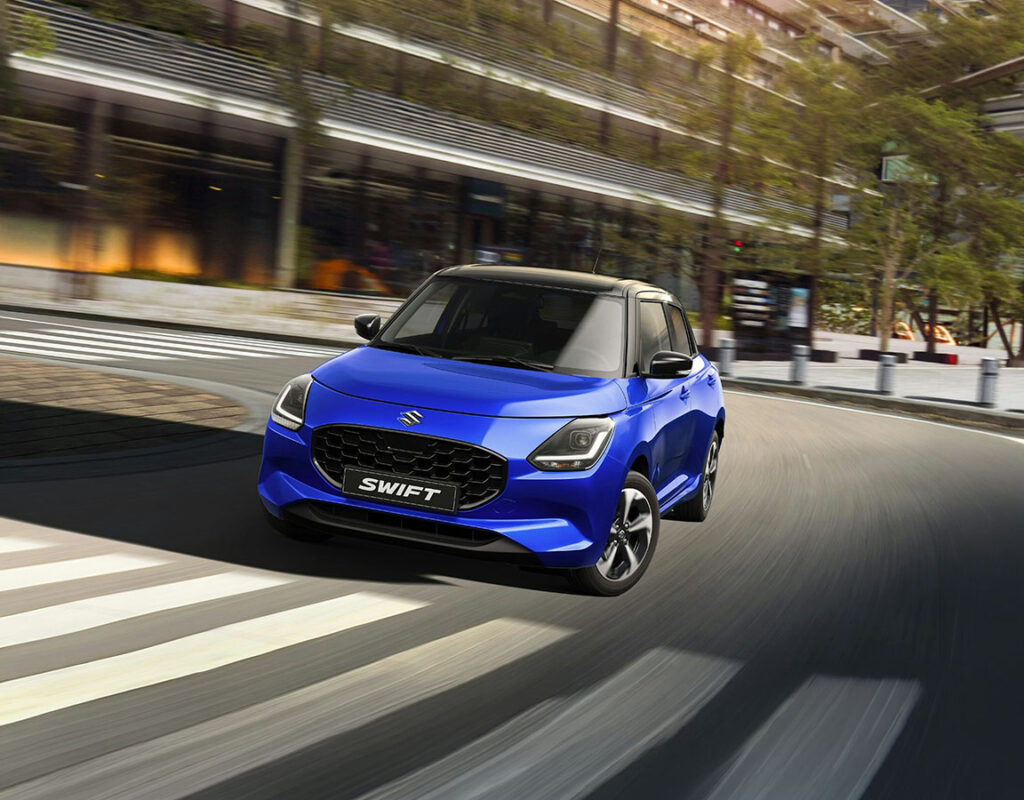 Ein blauer Suzuki Swift, präsentiert bei der Frey Auto AG, fährt auf einer kurvigen Stadtstraße und steht symbolisch für unsere Kompetenz in den Bereichen Verkauf, Service und Karosseriearbeiten rund um Suzuki-Fahrzeuge.