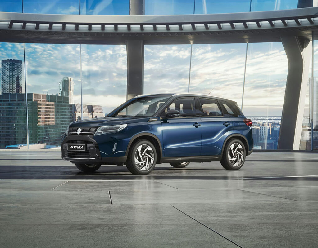 Ein blauer Suzuki Vitara bei der Frey Auto AG, ausgestellt zum Verkauf auf einem polierten Betonboden in unserem modernen, verglasten Showroom, der fachkundige Dienstleistungen für Toyota, Lexus und Suzuki anbietet, darunter MFK-Prüfung, Reifenservice, Wartung und Karosseriearbeiten – mit Blick auf die Skyline der Stadt.