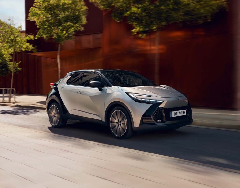 Ein silber-schwarzer Toyota C-HR, der bei der Frey Auto AG zum Verkauf steht, fährt durch eine moderne Stadtstraße, in der wir professionellen Service, MFK-Prüfung, Reifenservice, Karosseriearbeiten und Autowäsche anbieten.