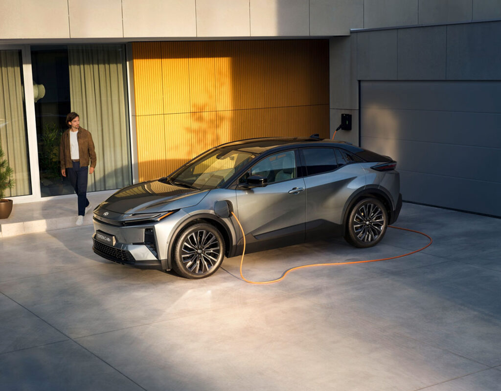 Ein graues Elektrofahrzeug vom Typ Toyota bZ4X der Firma Frey Auto AG, das in einer Einfahrt vor einem Haus geparkt und an eine Wandladestation angeschlossen ist, daneben steht ein Mann – ein Symbol für unsere kompetente Toyota-Verkaufsberatung und unseren professionellen Service für Elektrofahrzeuge.
