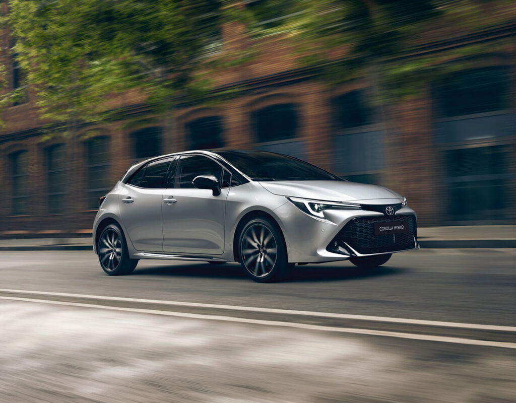 Ein silberner Toyota Corolla Hybrid fährt auf einer Stadtstraße – ein Symbol für den kompetenten Autoverkauf und die professionellen Wartungsdienstleistungen der Frey Auto AG, darunter die MFK-Prüfung und der Reifenservice.