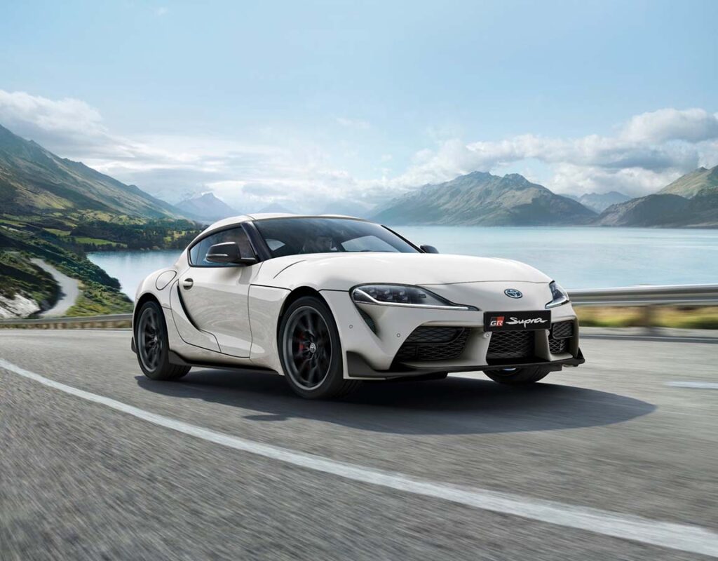 Ein weißer Toyota GR Supra fährt auf einer Küstenstraße in der Nähe von Bergen und einem See – ein Beispiel für das Premium-Fahrzeugangebot und die fachliche Kompetenz der Frey Auto AG, die unter anderem den Verkauf und die Wartung von Fahrzeugen der Marken Toyota, Lexus und Suzuki umfasst.