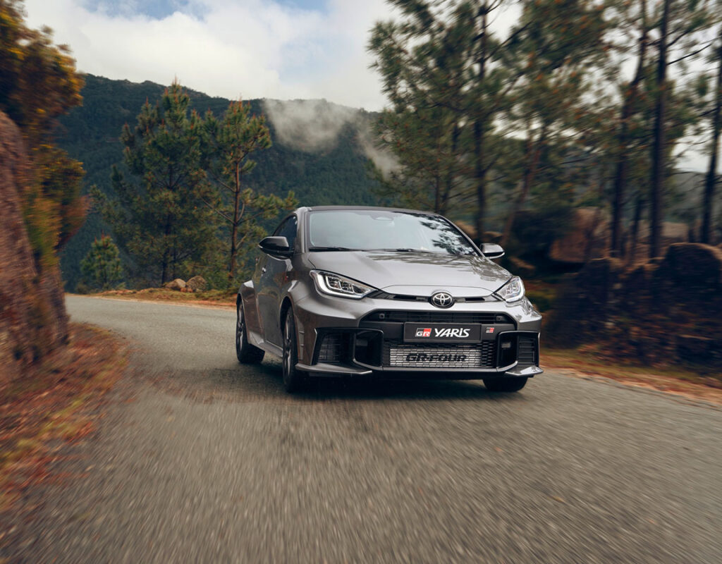 Ein grauer Toyota GR Yaris von Frey Auto AG fährt auf einer kurvenreichen Straße durch einen Wald und symbolisiert unseren Verkauf von Premium-Fahrzeugen sowie unsere fachkundigen Wartungsdienstleistungen, darunter MFK-Prüfung, Reifenservice und Karosseriearbeiten.