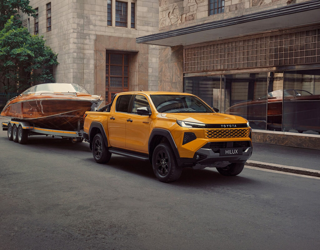 Ein gelber Toyota Hilux Pickup der Firma Frey Auto AG, der ein Motorboot aus Holz auf einem Anhänger zieht, steht auf unserem Gelände für professionelle Fahrzeugwartung, MFK-Prüfung und Autoverkauf.