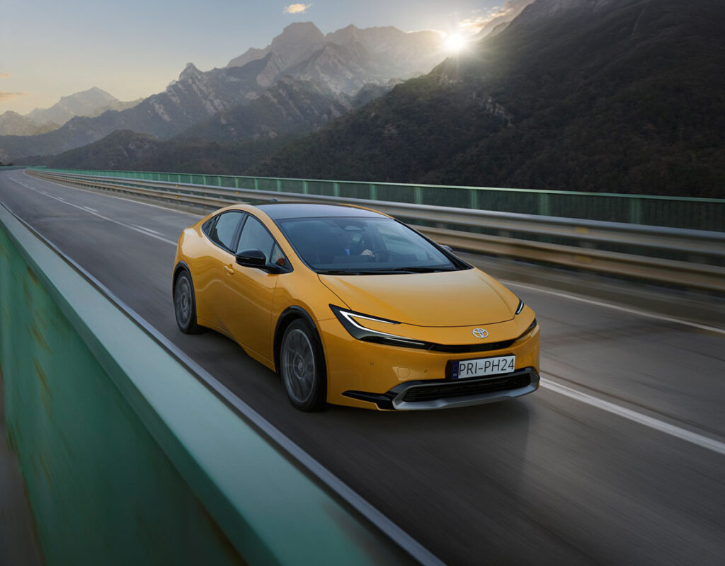 Ein gelber Toyota Prius der Frey Auto AG fährt auf einer Brücke durch eine bergige Landschaft und symbolisiert unsere Fachkompetenz im Autoverkauf und in der Fahrzeugwartung.