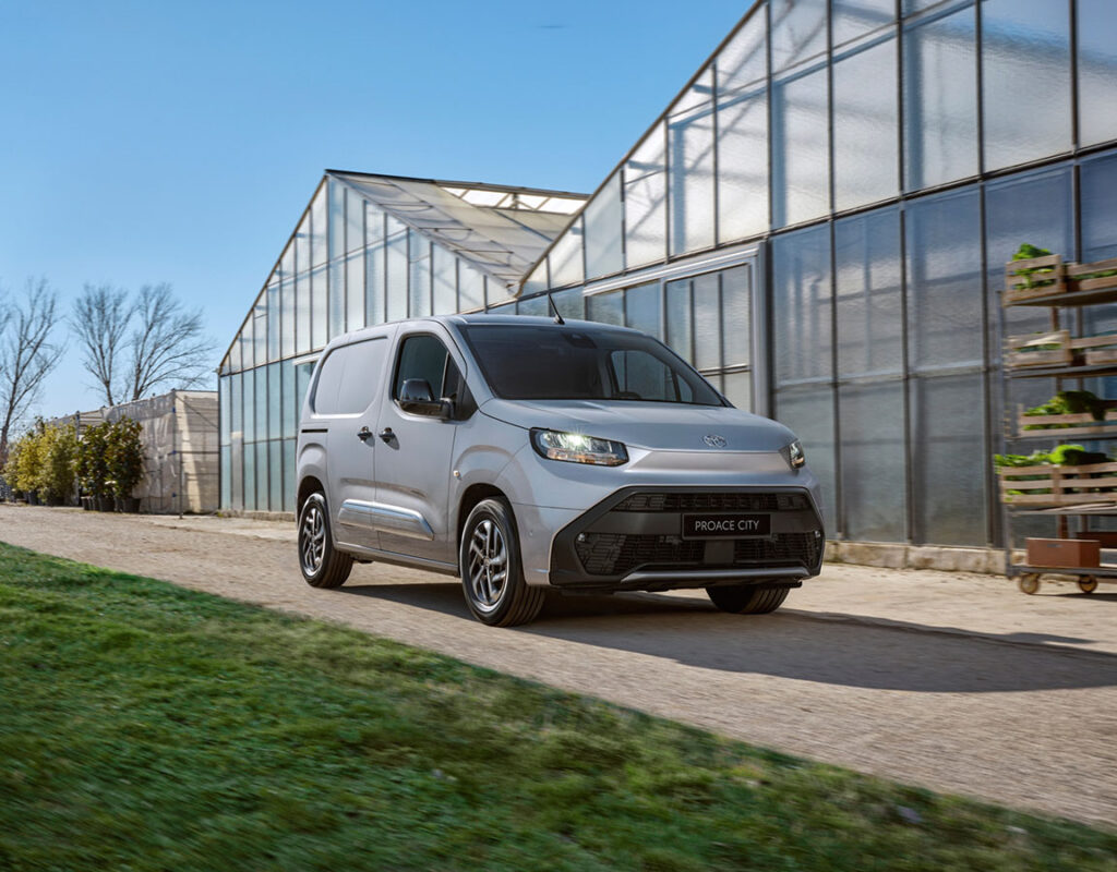 Ein silberner Toyota Proace City Van der Frey Auto AG fährt auf einem Weg neben einem Gewächshaus und symbolisiert unsere professionellen Kfz-Dienstleistungen, darunter Autoverkauf, MFK-Prüfung, Reifenservice, Wartung, Karosseriearbeiten und Autowäsche.