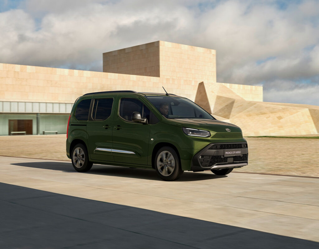 Ein grüner Toyota Proace City Verso fährt auf einer Betonstraße vor dem modernen Gebäude des Autohauses Frey Auto AG, das unser professionelles Autohaus und unser Kfz-Servicezentrum repräsentiert.