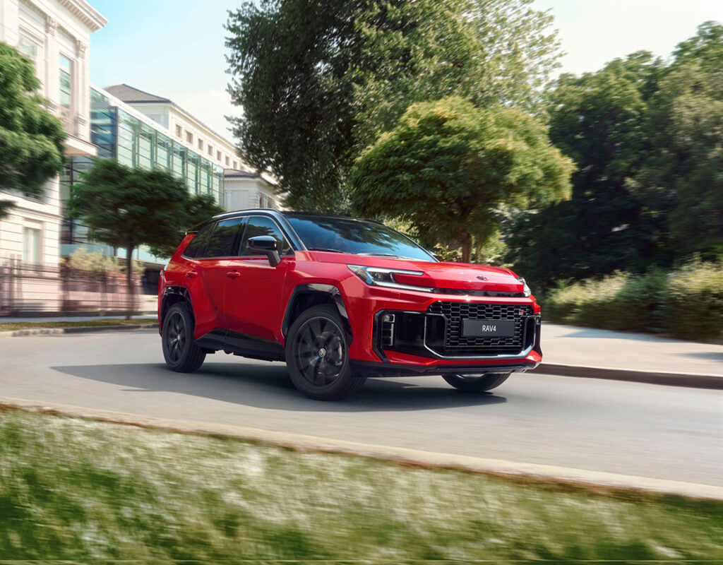 Ein roter Toyota RAV4 fährt eine von Bäumen und Gebäuden gesäumte Stadtstraße entlang – ein Symbol für das umfassende Angebot der Frey Auto AG in den Bereichen Autoverkauf, Fahrzeugservice und fachgerechte Karosseriearbeiten für Modelle der Marken Toyota, Lexus und Suzuki.