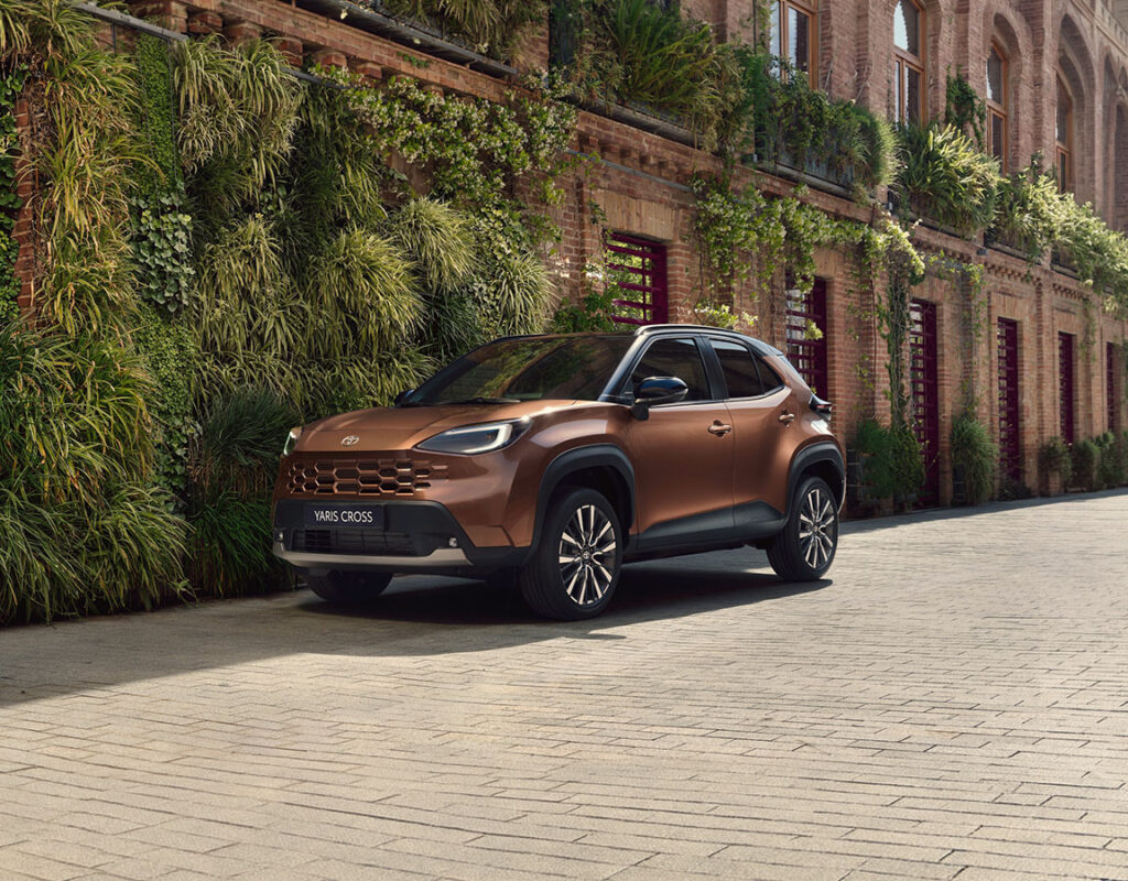 Ein brauner Toyota Yaris Cross steht auf einem Steinweg neben einem Backsteingebäude der Frey Auto AG mit einem vertikalen Garten – unserem führenden Zentrum für Autoverkauf, Wartung, Karosseriereparaturen und MFK-Prüfungen.
